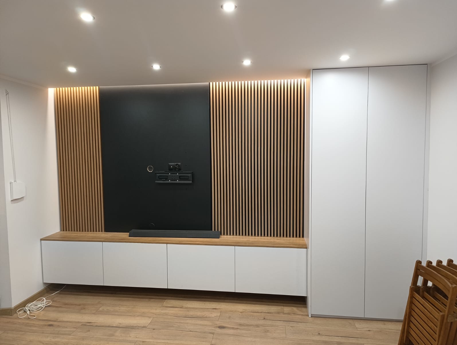 Nowoczesna ściana telewizyjna z drewnianymi lamelami, czarnym panelem na telewizor i białą szafką RTV oraz wysoką białą szafą w minimalistycznym stylu.