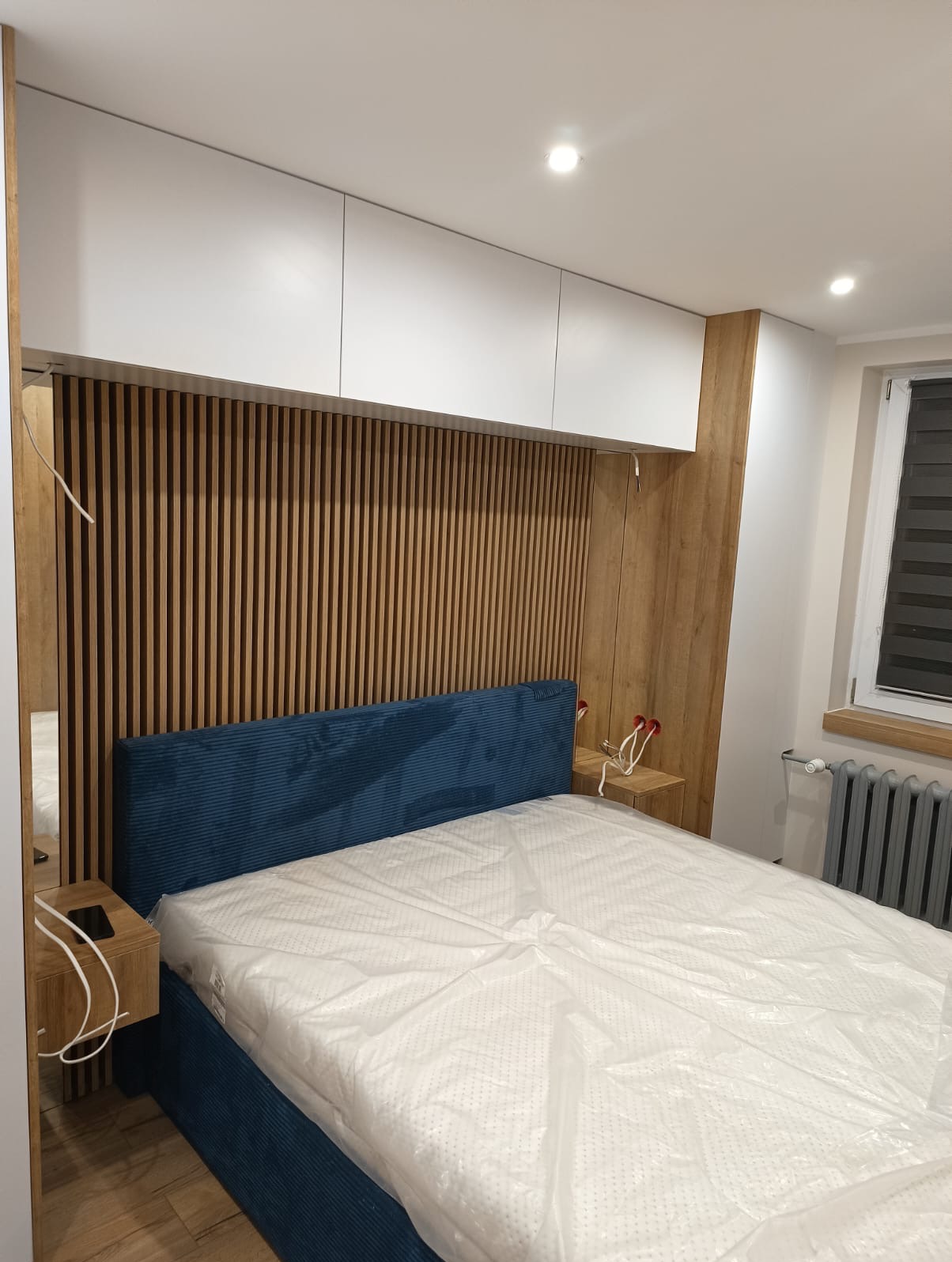 Nowoczesna sypialnia z łóżkiem z niebieskim wezgłowiem, drewnianą ścianką z pionowych listew, białymi szafkami wiszącymi i drewnianymi szafkami nocnymi, częściowo zakrytym materacem folią ochronną.