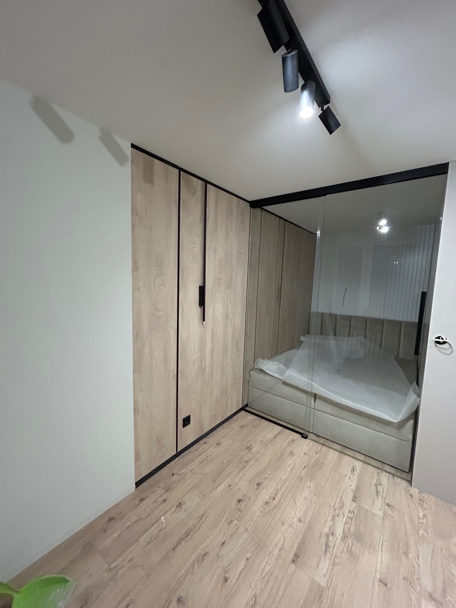 Nowoczesna szafa wnękowa z drewnopodobnymi frontami i czarnymi detalami, przylegająca do szklanej ściany oddzielającej sypialnię z łóżkiem przykrytym folią ochronną.