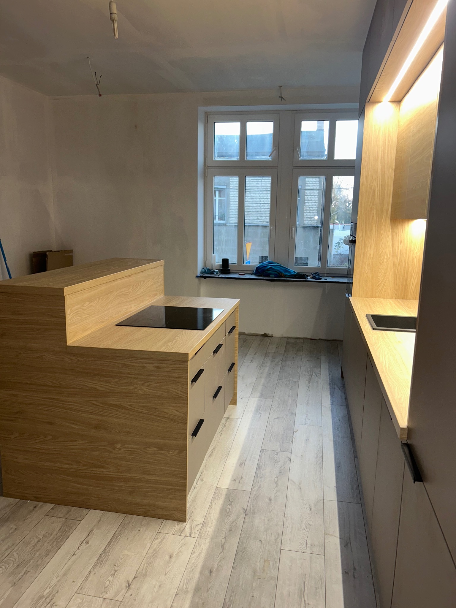 Wnętrze kuchni w trakcie montażu: drewniana wyspa z płytą indukcyjną i szufladami, zabudowa kuchenna z oświetleniem LED, surowa ściana, widoczne przewody elektryczne, okno z widokiem na zewnątrz.