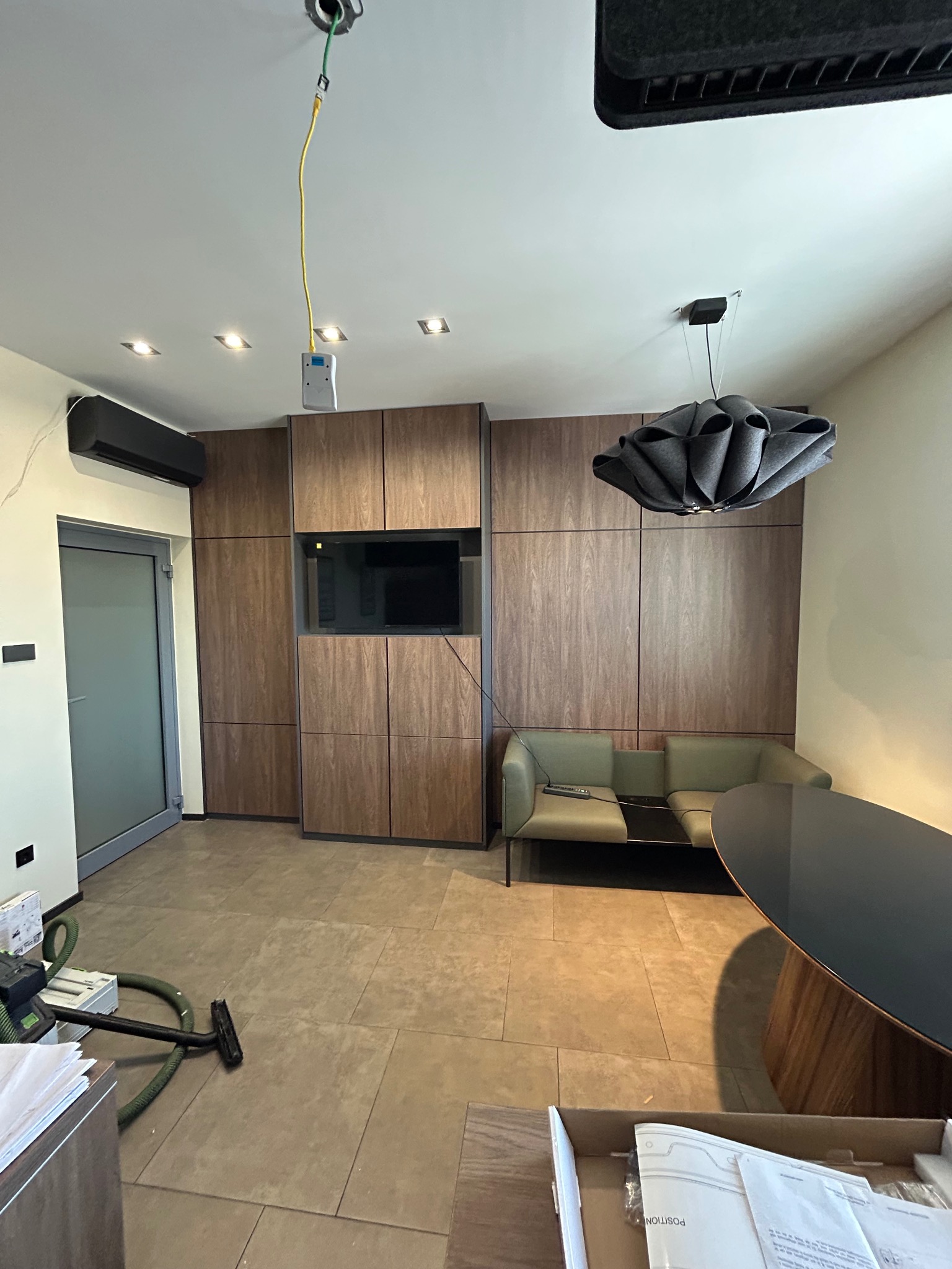 Nowoczesne wnętrze z zabudową meblową z drewnopodobnej płyty, telewizorem wkomponowanym w szafę, szarą sofą, designerską lampą i okrągłym stołem. Na podłodze leży odkurzacz przemysłowy, a z sufitu...