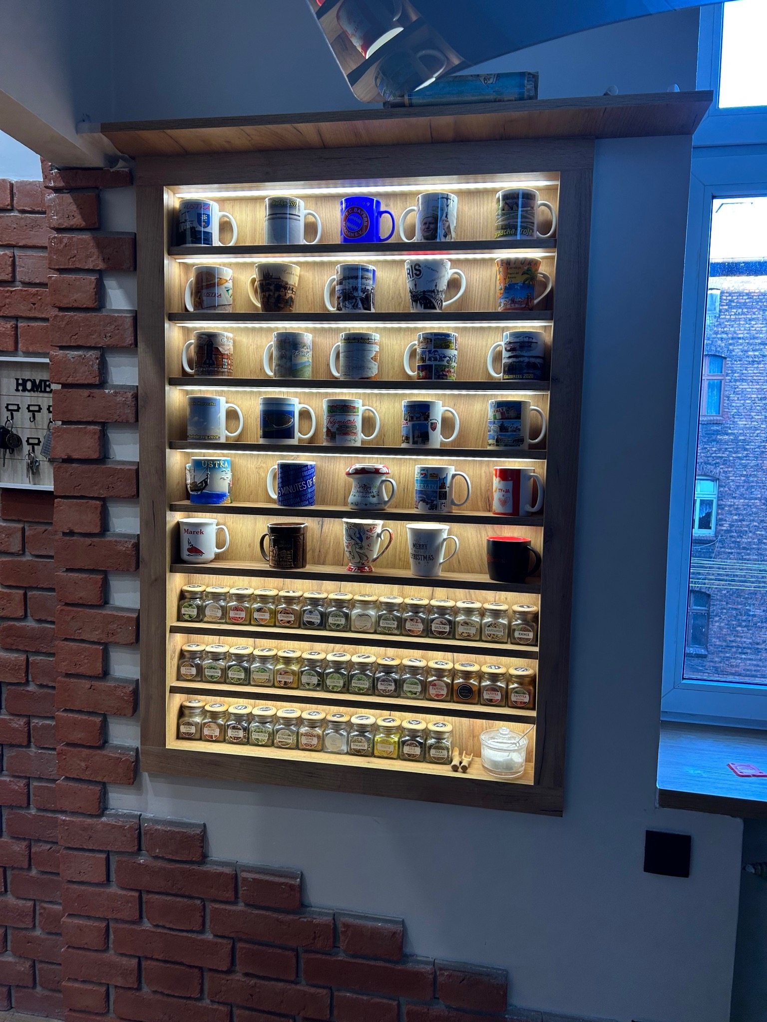 Półka z oświetleniem LED, wypełniona kolekcją kubków z motywami turystycznymi i słoiczkami z przyprawami, zamontowana na ścianie z cegły, obok okna.