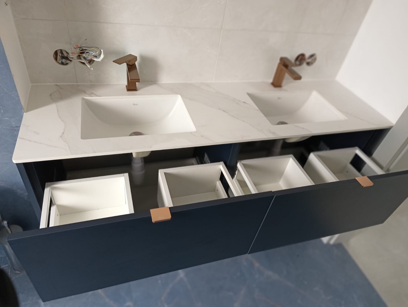 Podwójna umywalka z marmurowym blatem i otwartymi szufladami w szafce w kolorze granatowym; widoczne miedziane baterie i instalacja elektryczna w ścianie.
