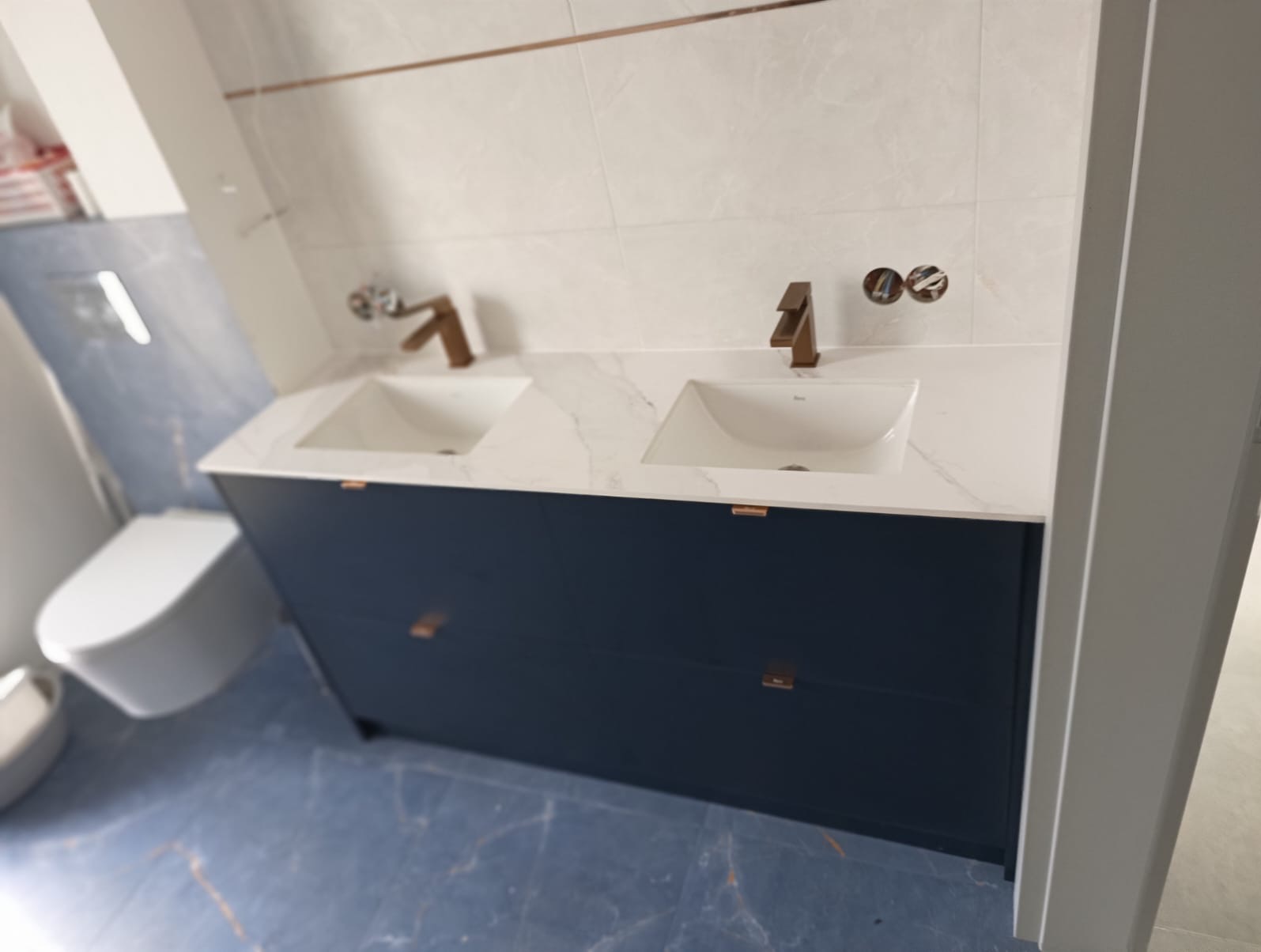 Nowoczesna łazienka z podwójną umywalką nablatową, marmurowym blatem i szafką w kolorze granatowym, uzupełniona o miedziane akcenty w postaci armatury i listew dekoracyjnych.