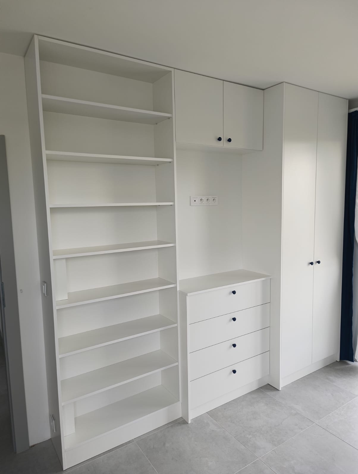 Biała zabudowa meblowa z otwartym regałem, szafkami wiszącymi i komodą z czterema szufladami, uzupełniona o szafę z dwoma drzwiami i ciemnymi, okrągłymi uchwytami.