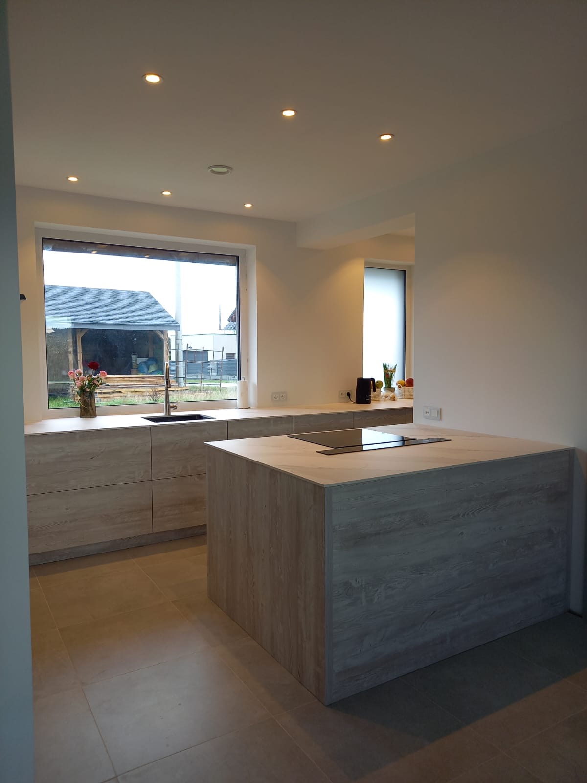 Nowoczesna kuchnia z wyspą i drewnopodobnymi frontami, widok na ogród przez duże okno, minimalistyczne oświetlenie punktowe.