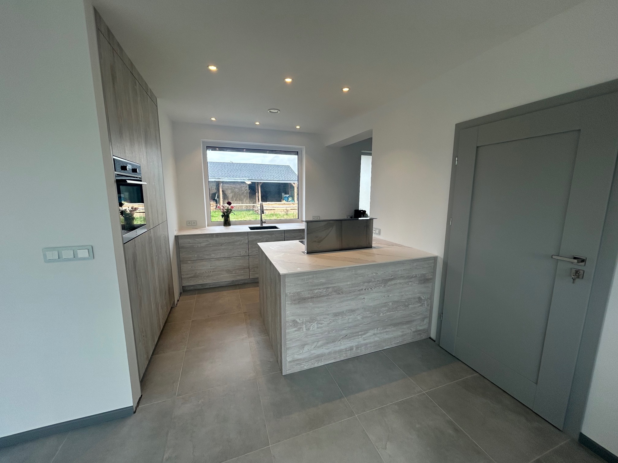 Nowoczesna kuchnia z wyspą w odcieniach szarości i beżu, widoczny piekarnik w zabudowie i okno z widokiem na ogród.