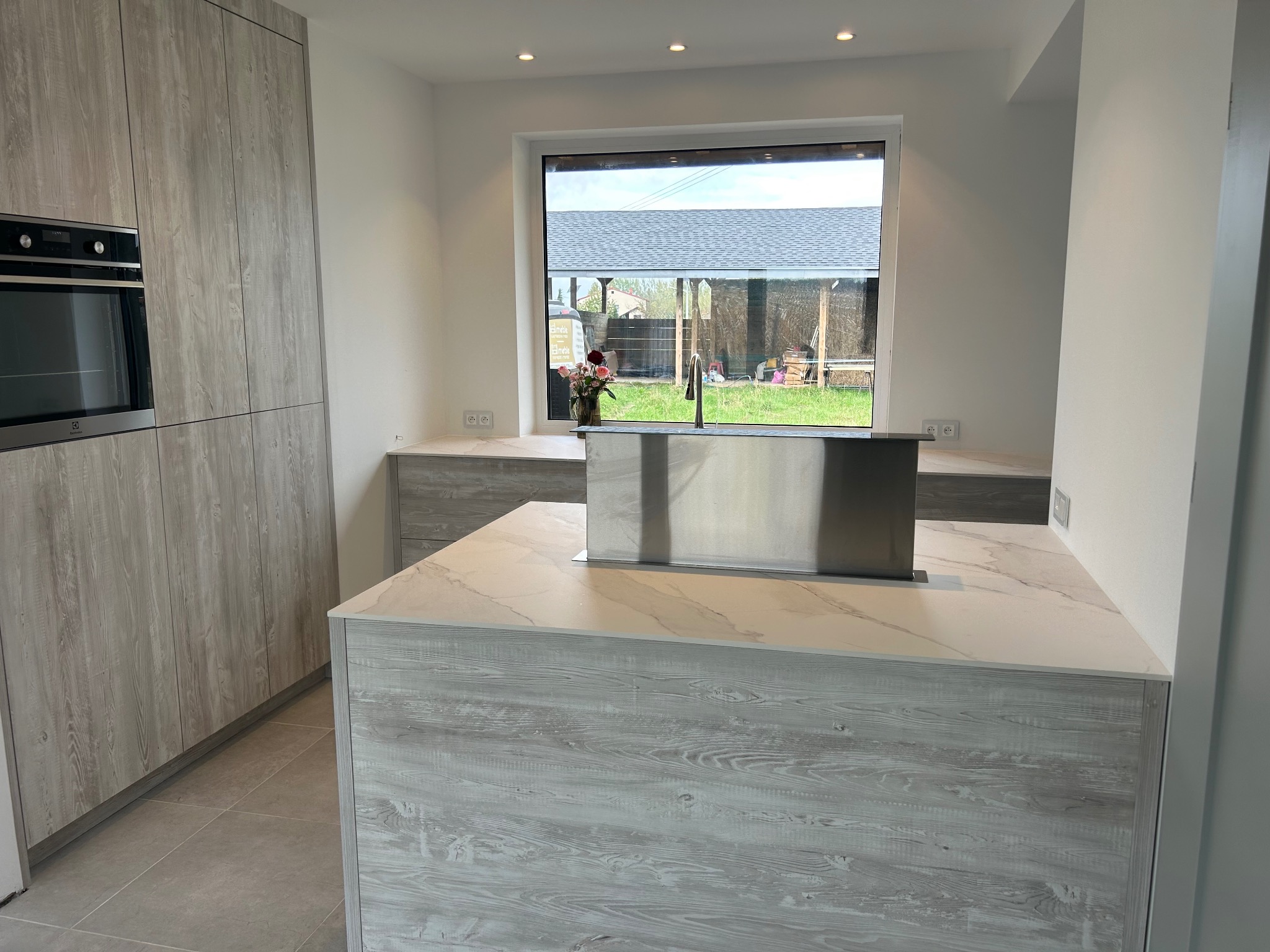 Nowoczesna kuchnia z wyspą i wyciągiem blatowym, fronty szafek w odcieniach szarości z wyraźnym rysunkiem drewna, duży panoramiczny widok na ogród przez okno.