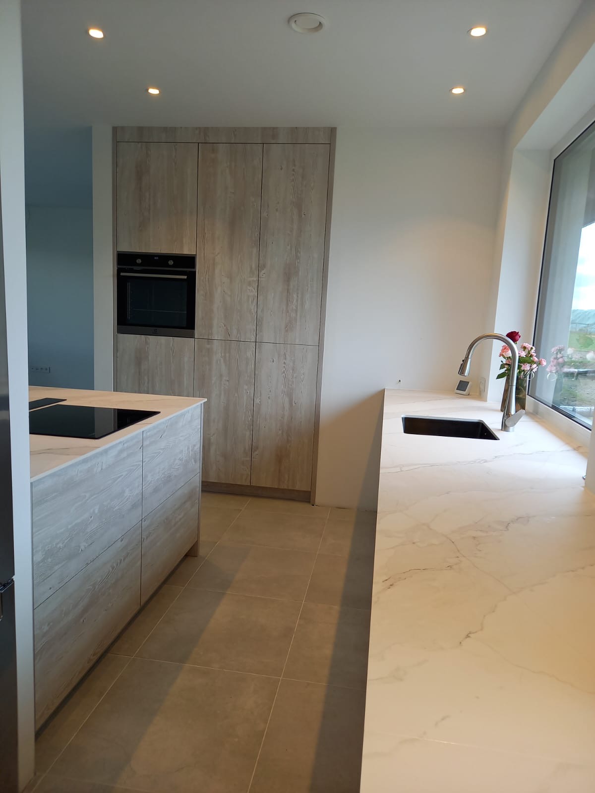 Nowoczesna kuchnia z wyspą w odcieniach beżu i bieli, z drewnopodobnymi frontami i marmurowym blatem, oświetlona punktowymi lampami sufitowymi, z widokiem na zieleń za oknem.