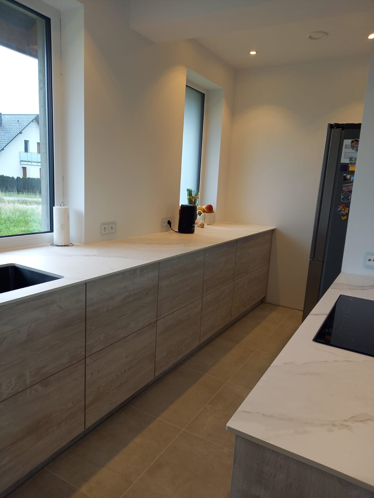 Nowoczesna kuchnia z drewnopodobnymi frontami szafek, jasnym blatem z marmurowym wzorem, oknem z widokiem na ogród i zabudowaną lodówką.