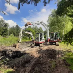 Dwa biało-czerwone mini-koparki Takeuchi na błotnistym terenie, prawdopodobnie w trakcie pogłębiania lub czyszczenia stawu, w tle widoczna zieleń drzew i błękitne niebo z chmurami.