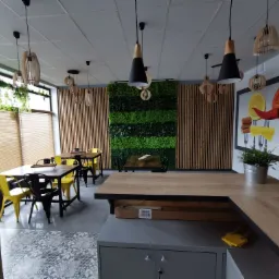 Nowoczesne wnętrze biura z drewnianymi panelami, zieloną ścianą z mchu, designerskimi lampami wiszącymi i stołami z żółtymi i czarnymi krzesłami, uzupełnione dekoracyjnym panelem z motywem warzyw...