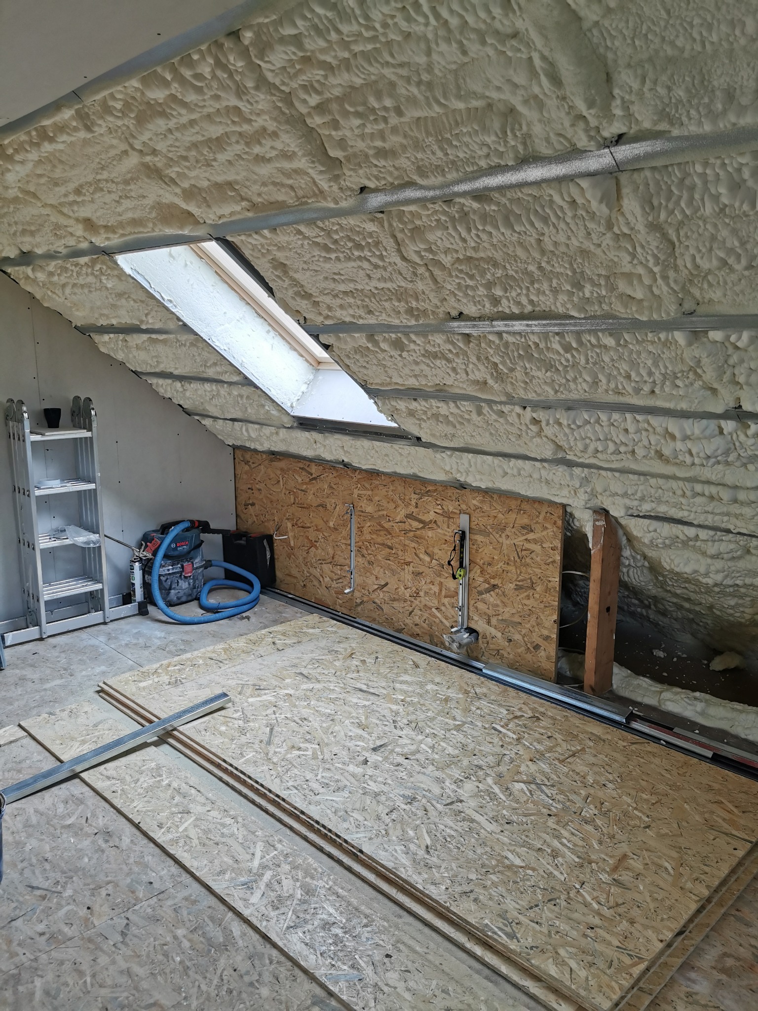 Poddasze w trakcie ocieplania pianą PUR, widoczne płyty OSB na podłodze, okno dachowe i drabina aluminiowa w rogu pomieszczenia.