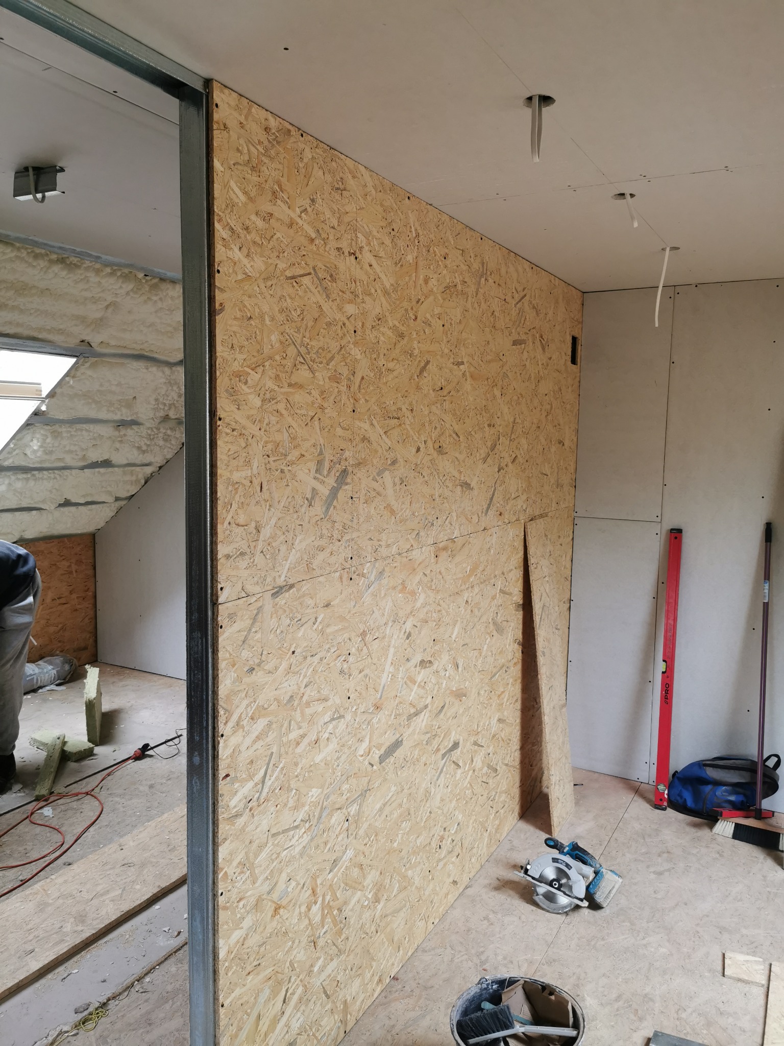 Montaż ściany z płyty OSB na metalowym stelażu w trakcie prac wykończeniowych poddasza, widoczna piła tarczowa, poziomica i fragment ocieplenia wełną mineralną.