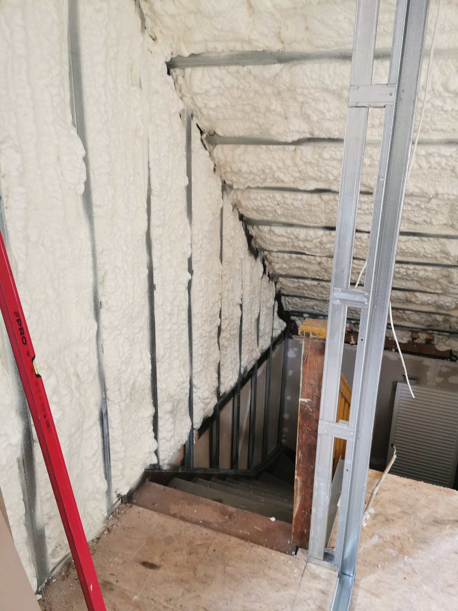 Aplikacja pianki izolacyjnej na poddaszu, widoczne metalowe profile konstrukcyjne, czerwona poziomica i fragment schodów.