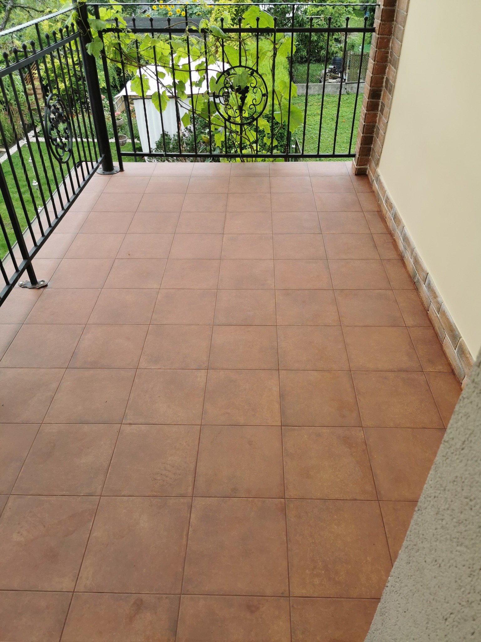 Wykafelkowany balkon z czarną, kutą balustradą, widok na zielony ogród z winoroślą.