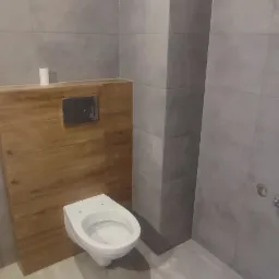 Nowoczesna toaleta podwieszana z drewnianą obudową stelaża, szare płytki na ścianach i podłodze, minimalistyczny design.