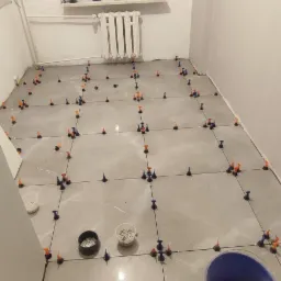 Szare płytki podłogowe w trakcie układania, z użyciem systemu poziomowania płytek, widoczne krzyżyki dystansowe, sznurek traserski, wiadro z akcesoriami i poziomica laserowa przy oknie.