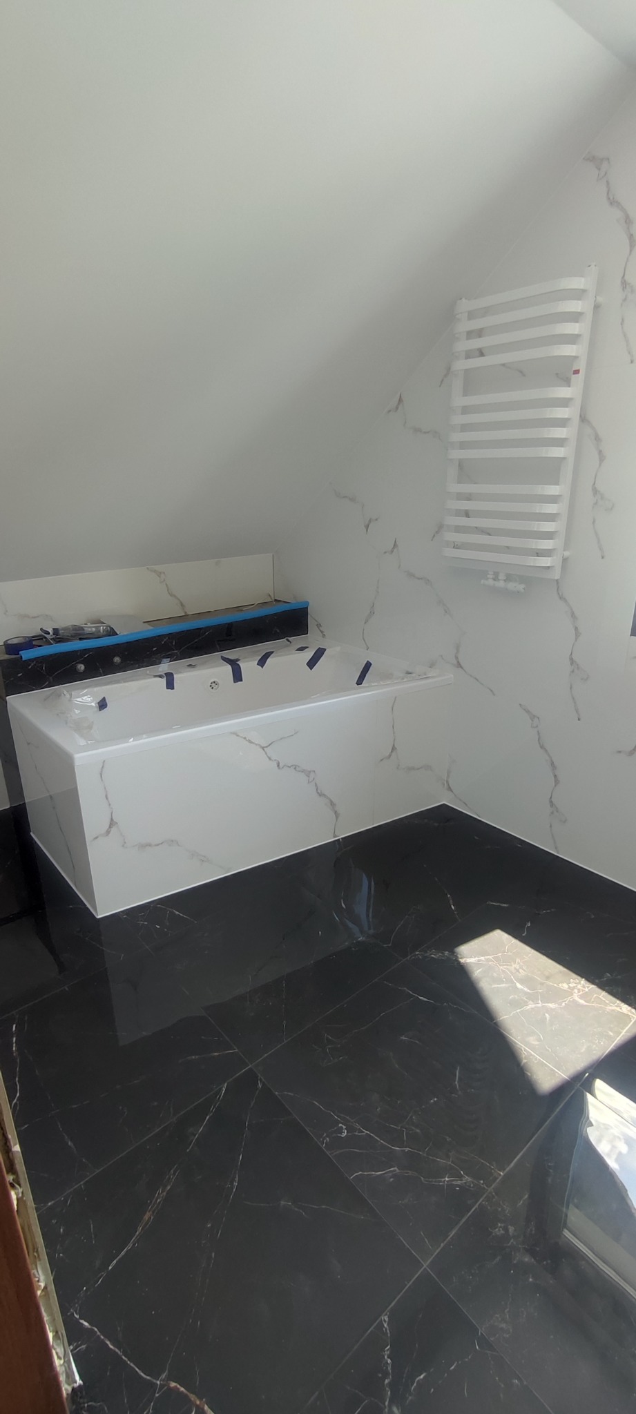 Biała wanna z hydromasażem w łazience z czarną marmurową podłogą i białymi marmurowymi ścianami, grzejnik drabinkowy na ścianie, folia ochronna na wannie.