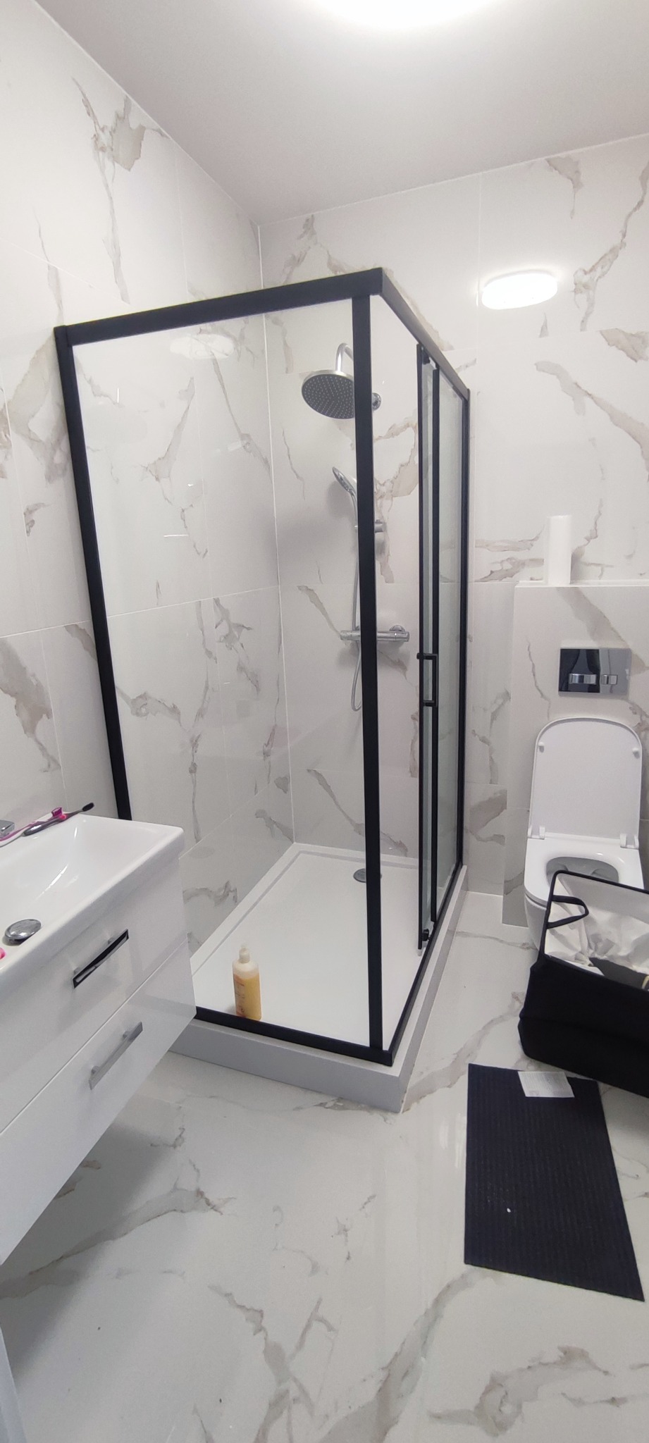 Nowoczesna kabina prysznicowa z czarną ramą na tle marmurowych płytek, widoczna umywalka z szafką i toaleta, na podłodze leży czarna mata.