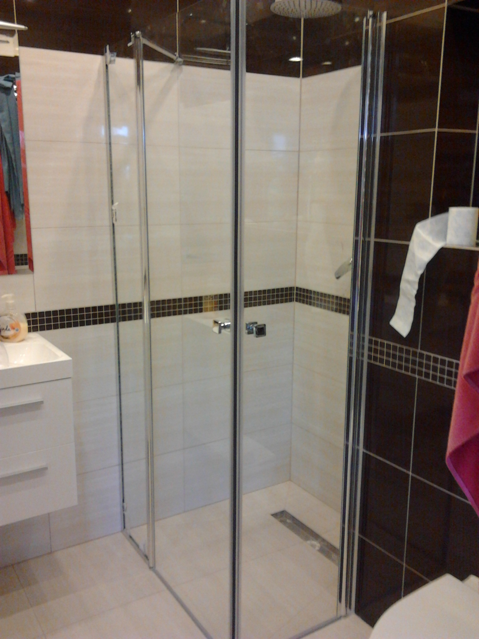 Nowoczesna kabina prysznicowa z deszczownicą, jasne płytki z dekoracyjnym pasem mozaiki, biała umywalka z szafką i dozownikiem mydła, ciemnobrązowe płytki na ścianie obok z uchwytem na papier...