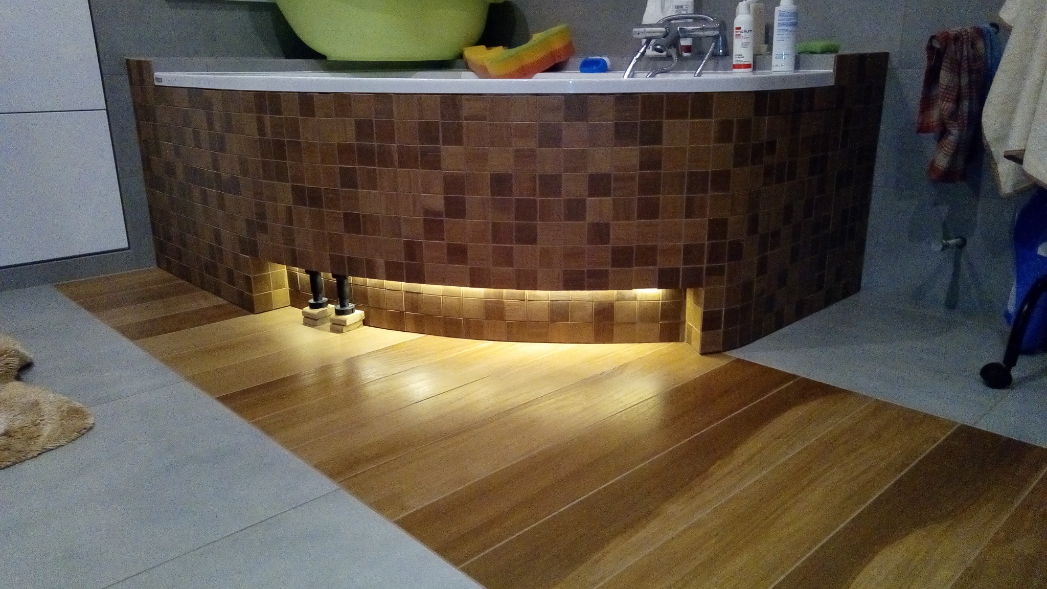 Obudowa wanny wykończona mozaiką z podświetleniem LED u dołu, drewnopodobne płytki na podłodze, widoczne rury odpływowe.