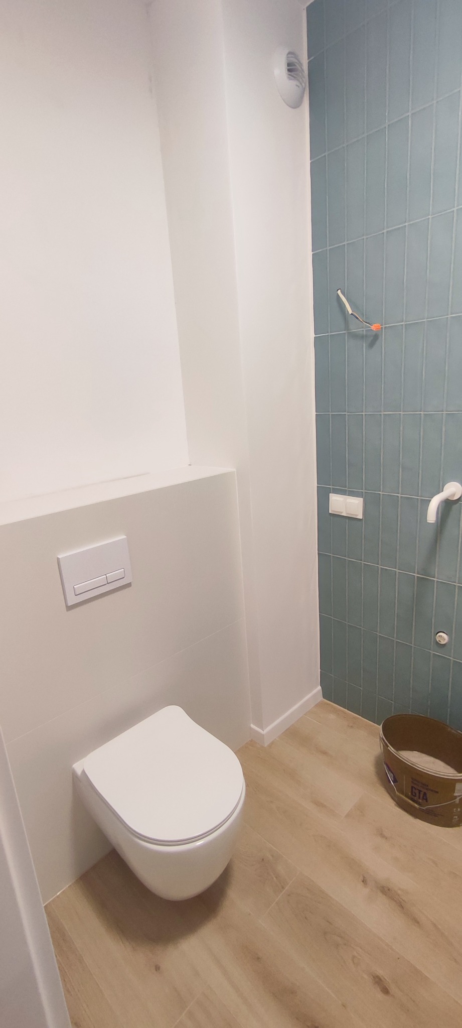 Nowoczesna łazienka z podwieszaną toaletą, turkusowymi płytkami na ścianie i drewnopodobną podłogą. Widoczny wystający kabel elektryczny i wiadro z zaprawą.