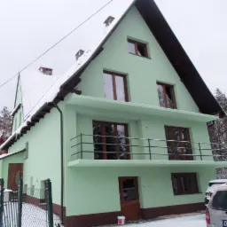 Świeżo pomalowany, dwupiętrowy dom z balkonem w pastelowej zieleni, z brązowymi akcentami, widziany z zewnątrz w zimowej scenerii z ośnieżonym dachem i samochodem na podjeździe.