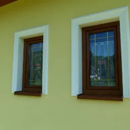 Dwa okna z brązowymi ramami i białymi obramowaniami na żółtej elewacji budynku, widoczne zasłony w oknach.