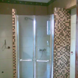 Szklana kabina prysznicowa z chromowanymi uchwytami, otoczona mozaiką z brązowo-beżowych płytek, widoczna słuchawka prysznicowa na ścianie wewnątrz kabiny, ujęcie z niskiego kąta.