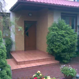 Wejście do domu z zadaszeniem, wyłożone terakotą, otoczone zielenią i kwiatami w donicach.