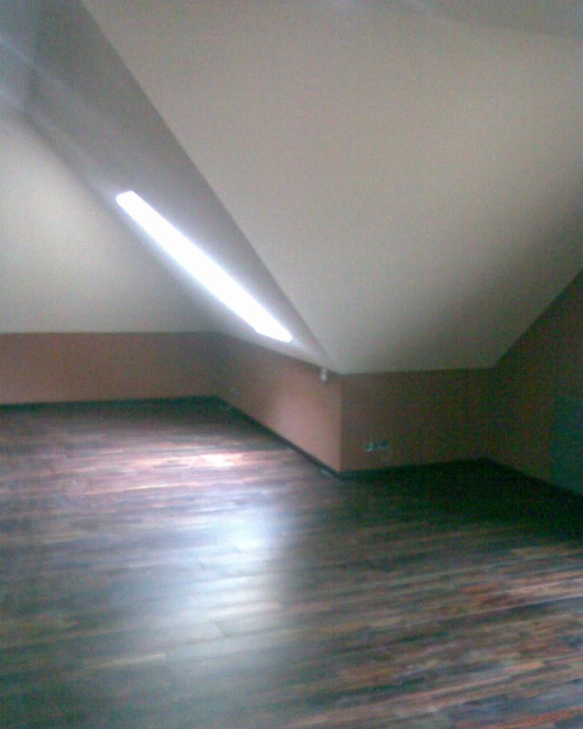 Poddasze po remoncie z brązową ścianą, skośnym sufitem i nową, ciemną podłogą z paneli.