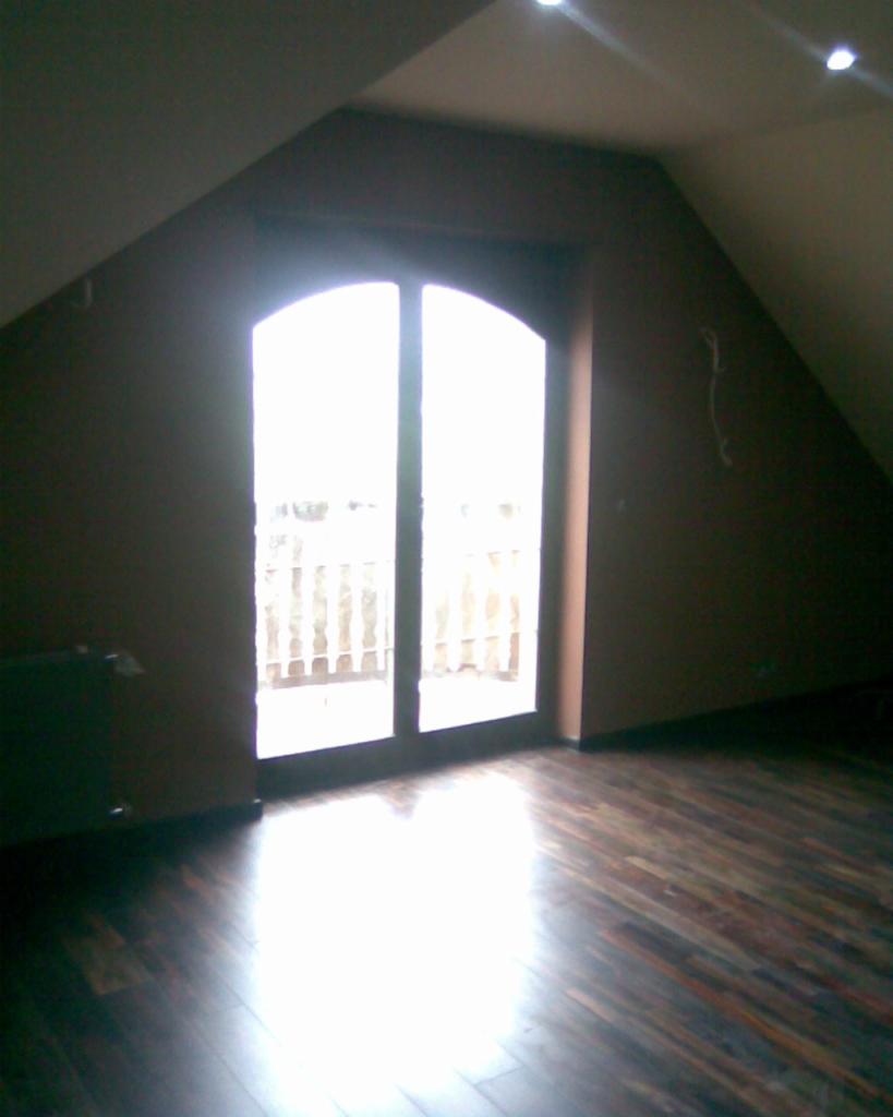 Poddasze z oknem balkonowym i parkietem, ściany pomalowane na brązowo, widoczne punkty świetlne w suficie.