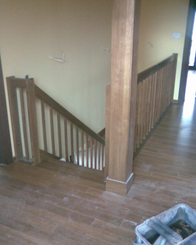 Drewniane schody z balustradą w trakcie montażu, widoczna fuga między płytkami podłogowymi i wiadro z zaprawą.