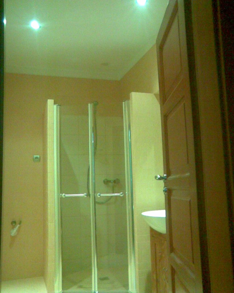 Jasna łazienka z kabiną prysznicową z przesuwanymi drzwiami, umywalką częściowo widoczną za otwartymi drewnianymi drzwiami, oraz beżowymi ścianami i podłogą.