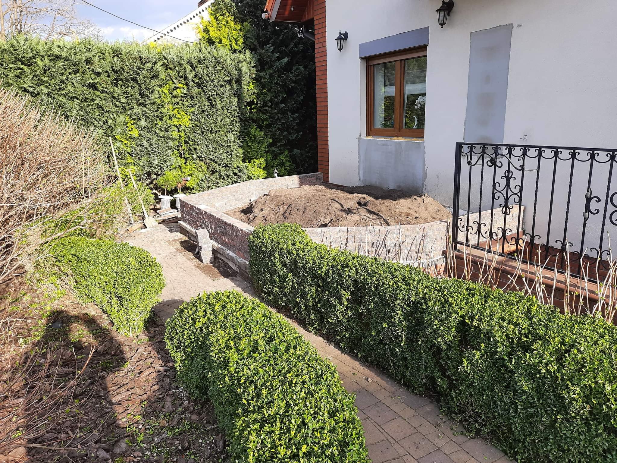 Przygotowanie pod taras: wykopana ziemia w obudowie z cegieł przy domu, widoczna ścieżka z kostki brukowej oraz ozdobne krzewy.