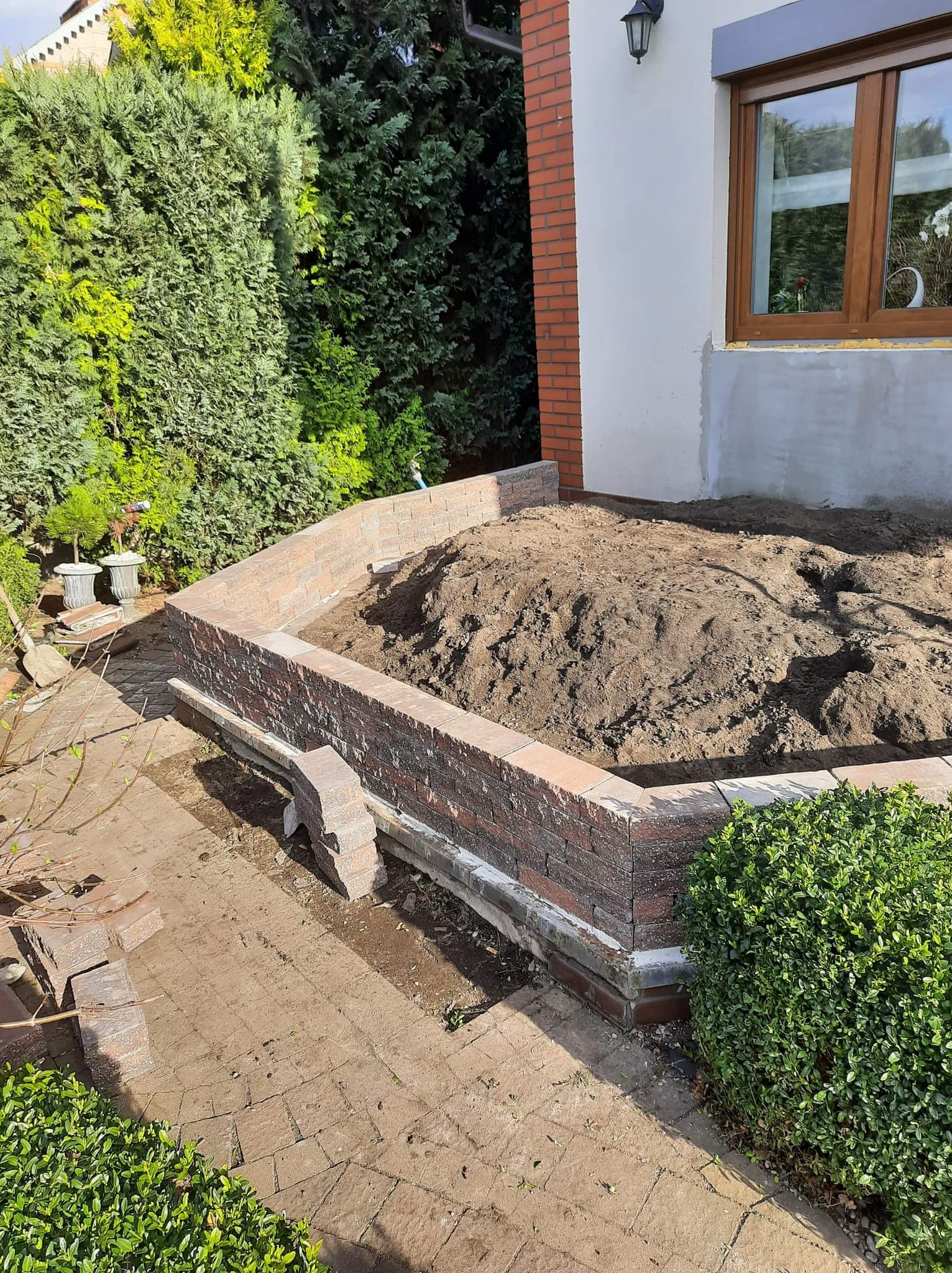 Budowa podwyższonej rabaty kwiatowej z cegieł klinkierowych, wypełnionej ziemią, z kostką brukową w tle i zielenią ogrodową w otoczeniu.