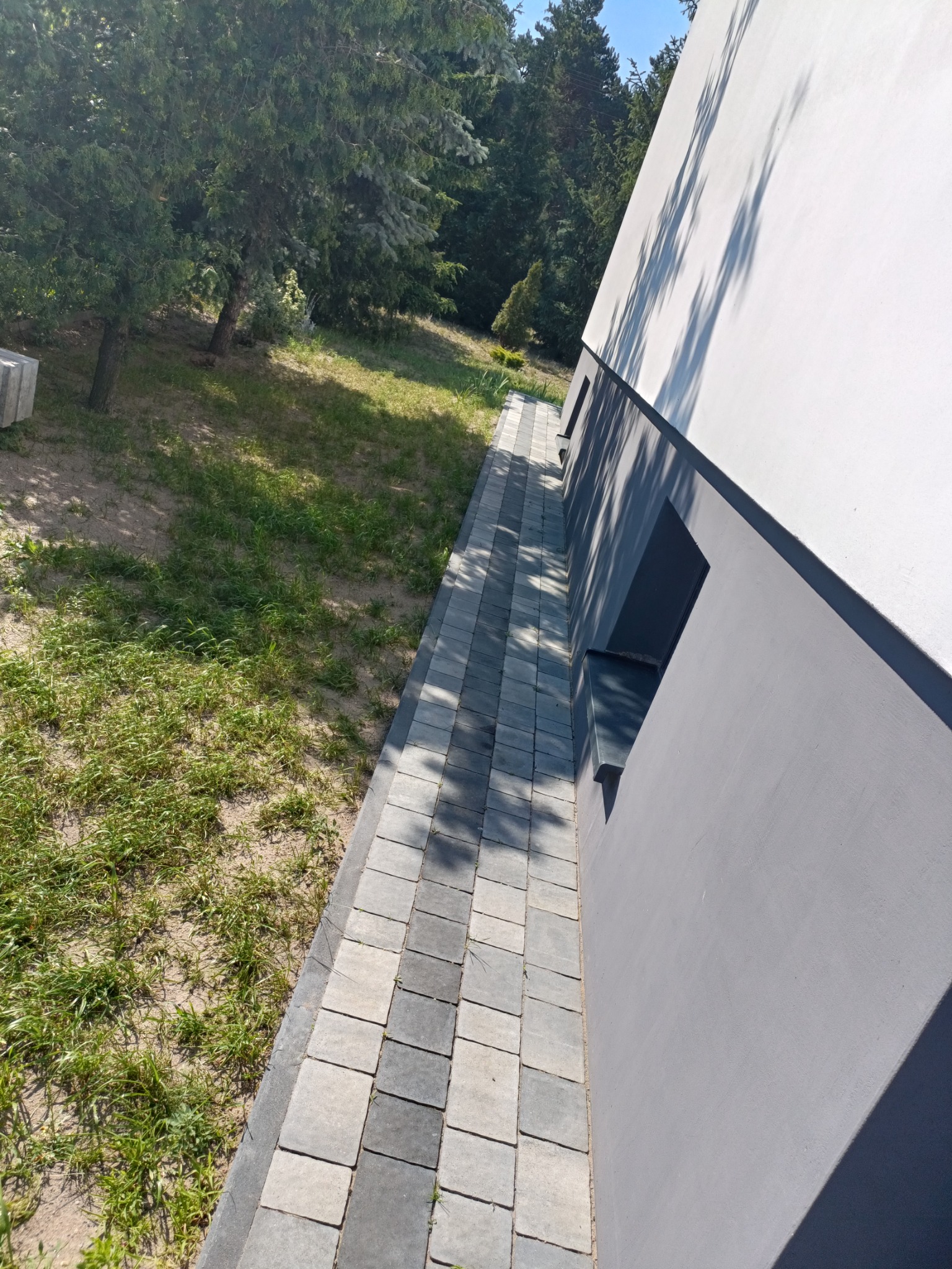 Ścieżka z kostki brukowej w odcieniach szarości, przylegająca do jasnej ściany budynku, z widoczną zielenią trawy i drzew w tle.