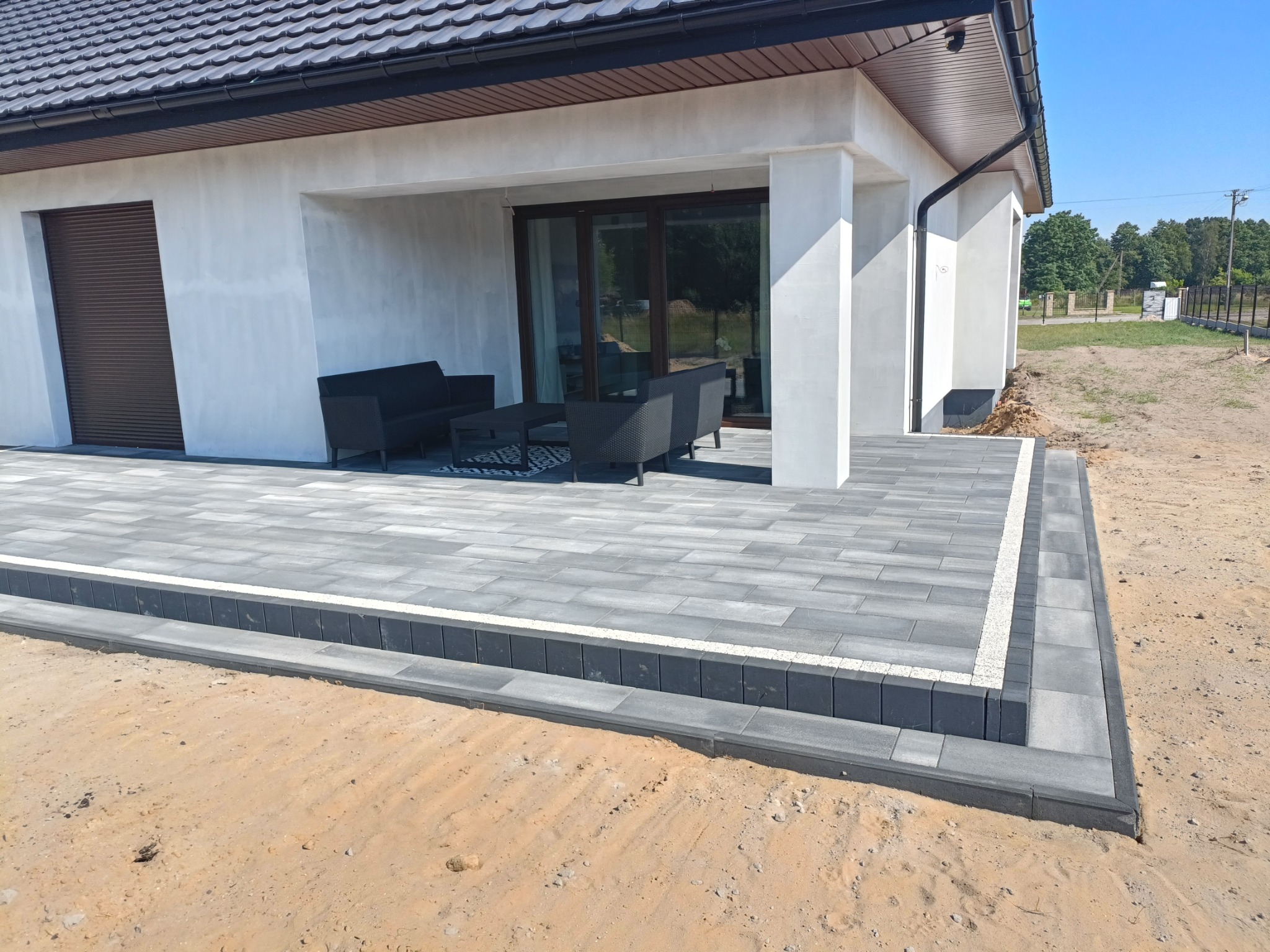 Nowoczesny taras z jasnoszarej kostki brukowej, obrzeża z ciemniejszej kostki, meble ogrodowe w kolorze antracytowym, dom w tle.