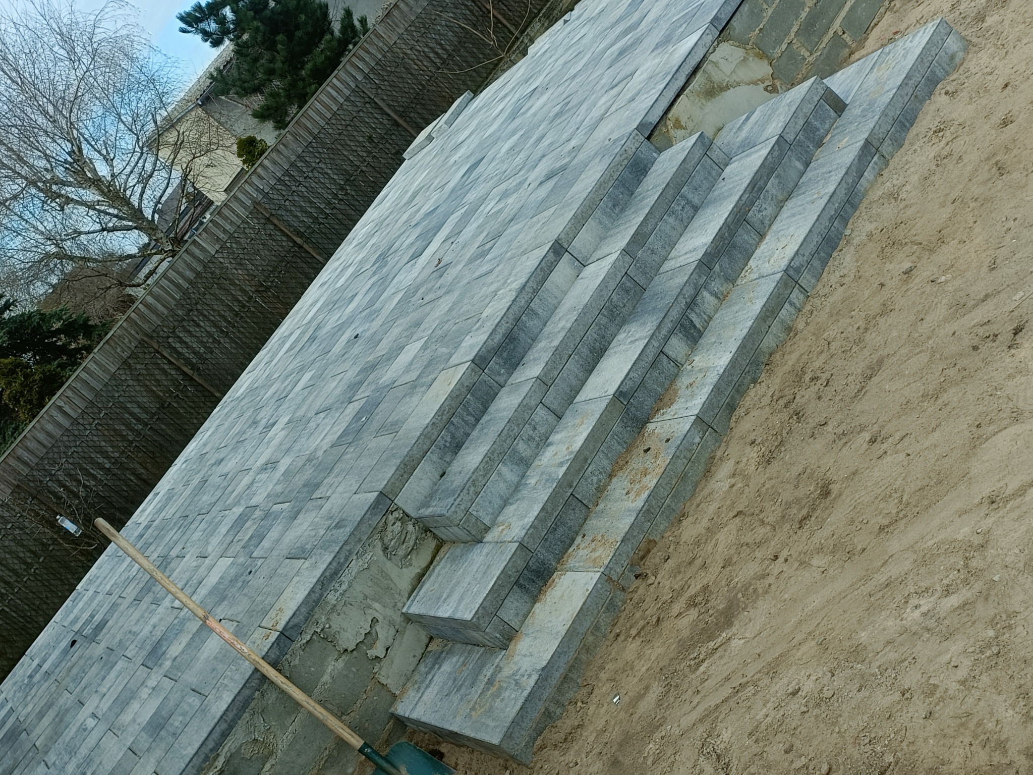 Kładzenie szarej kostki brukowej na tarasie, widoczne niedokończone schody i łopata.