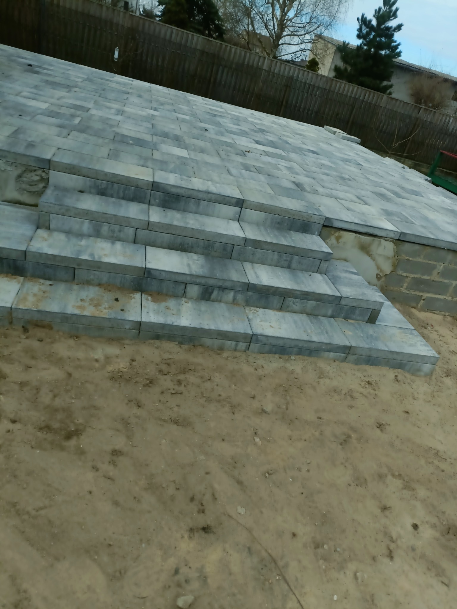 Niedokończone schody z szarej kostki brukowej prowadzące na podwyższony taras, widoczne bloczki fundamentowe i piasek.