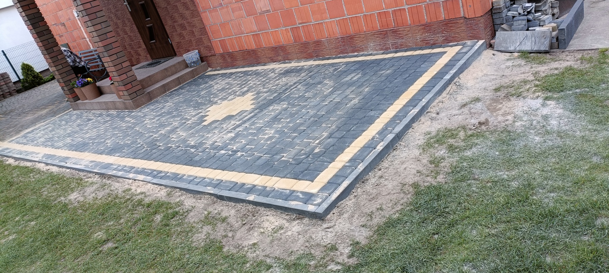 Ułożona kostka brukowa w kolorach szarym i żółtym przed wejściem do domu z cegły, wzór gwiazdy na środku, widoczne obrzeża i trawnik.