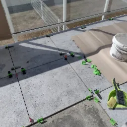 Układanie szarych płytek na balkonie z użyciem systemu poziomowania, widoczny kubeł z masą szpachlową 'Finish Plus', zmiotka z szufelką i gąbka do czyszczenia.