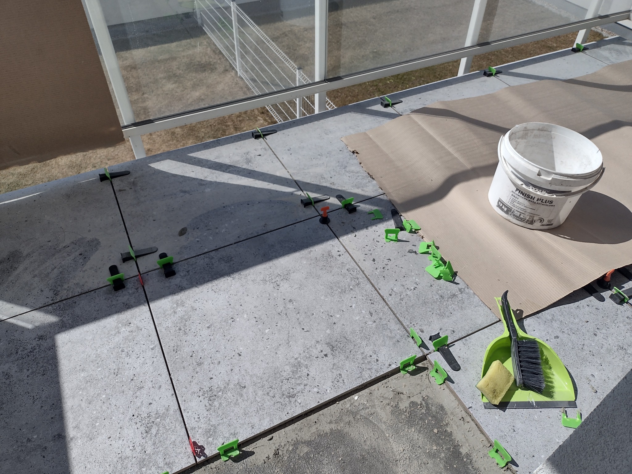 Układanie szarych płytek na balkonie z użyciem systemu poziomowania, widoczny kubeł z masą szpachlową 'Finish Plus', zmiotka z szufelką i gąbka do czyszczenia.