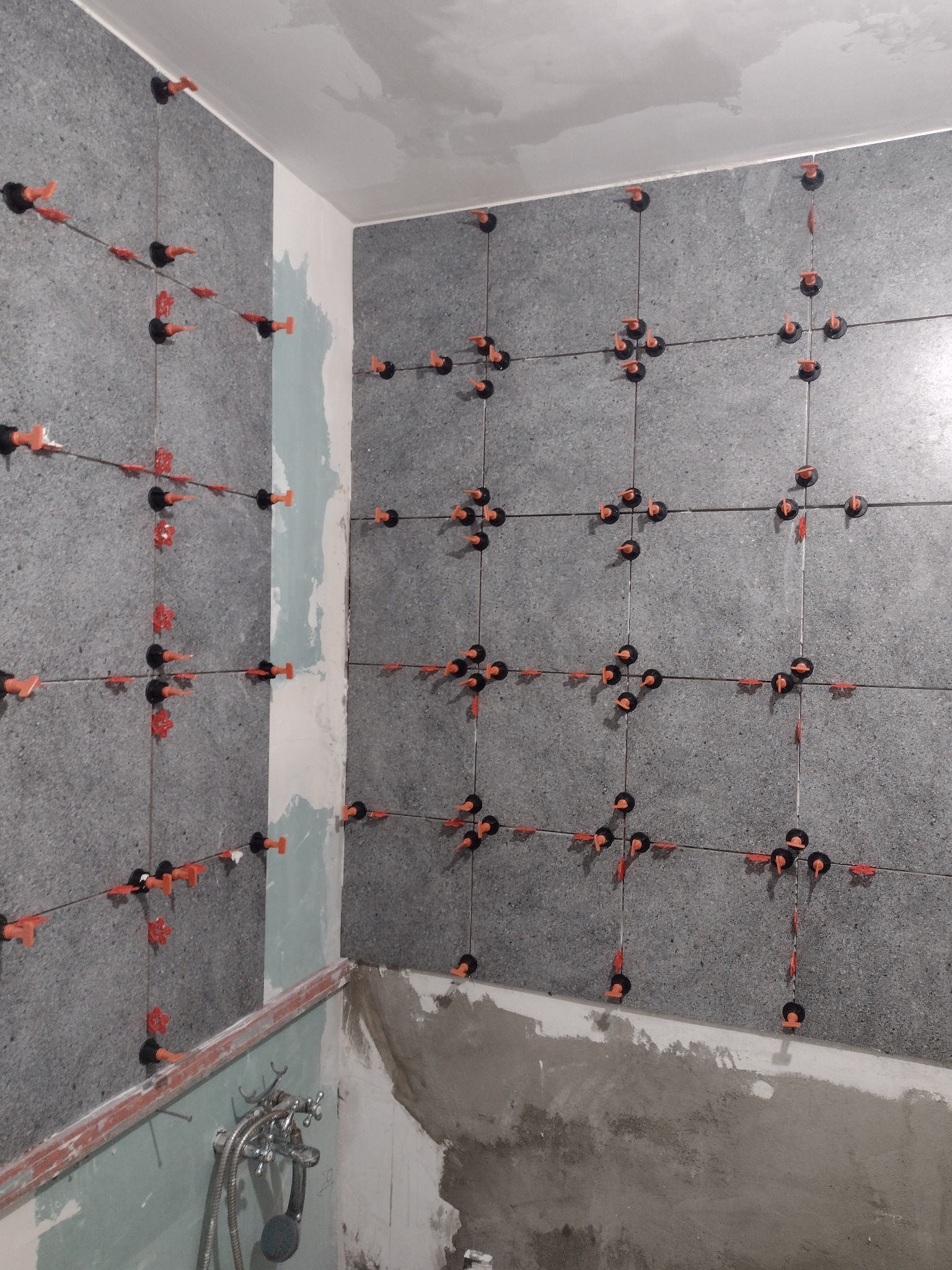 Świeżo ułożone, szare płytki ścienne z systemem poziomowania płytek (czerwone kliny i czarne klipsy) w trakcie prac wykończeniowych w łazience. Widoczna surowa ściana w dolnej części.