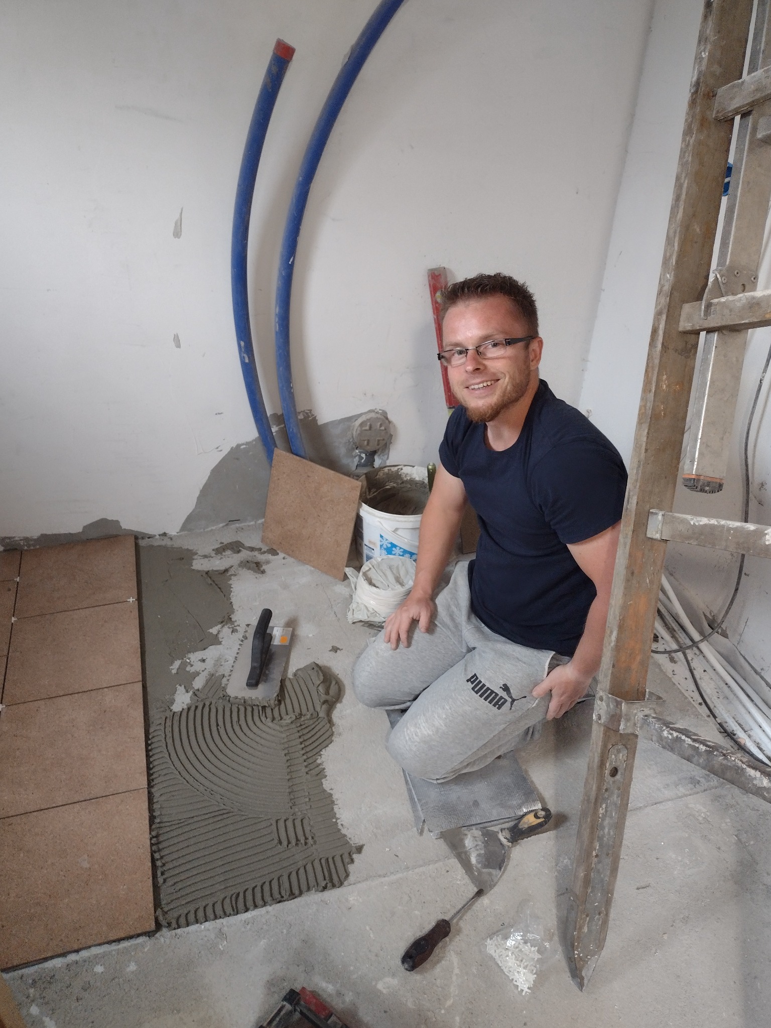 Uśmiechnięty glazurnik w okularach, klęczy obok świeżo ułożonych brązowych płytek podłogowych na warstwie kleju, widoczne narzędzia i drabina w tle.