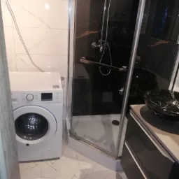 Nowoczesna łazienka z kabiną prysznicową typu walk-in z czarnymi marmurowymi ścianami, białą pralką i umywalką nablatową z czarnego marmuru.