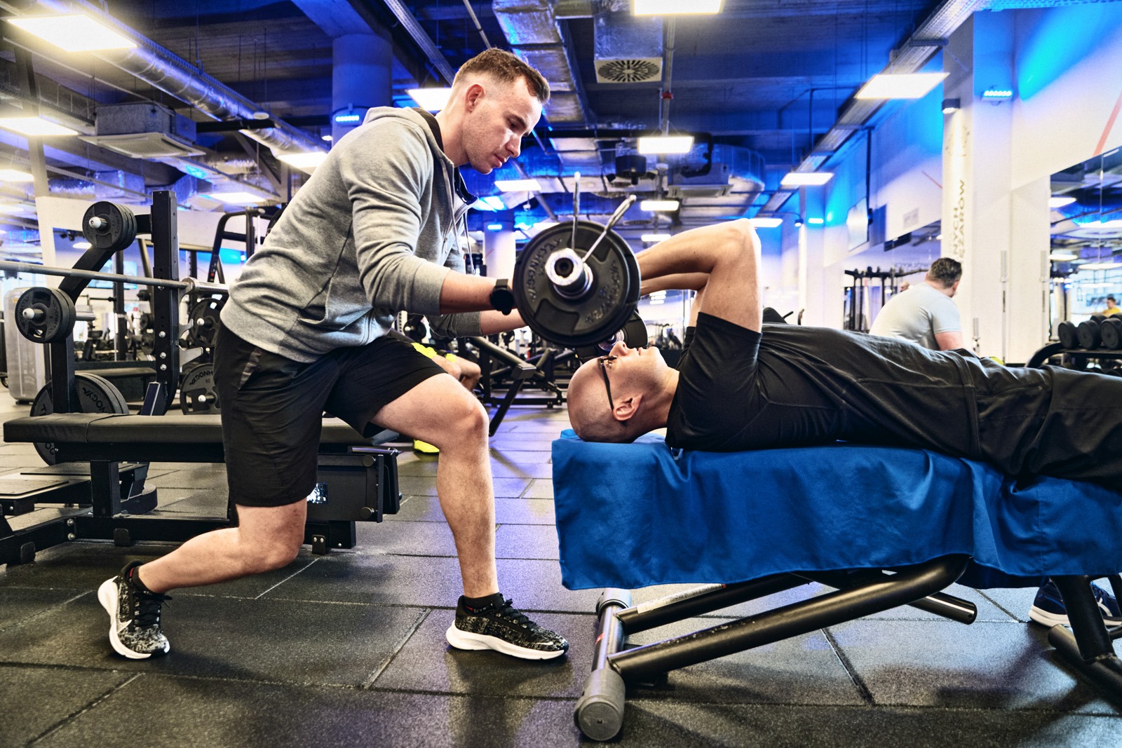 Trener personalny asekuruje mężczyznę leżącego na ławeczce podczas wyciskania sztangi w nowoczesnej siłowni z niebieskim oświetleniem.