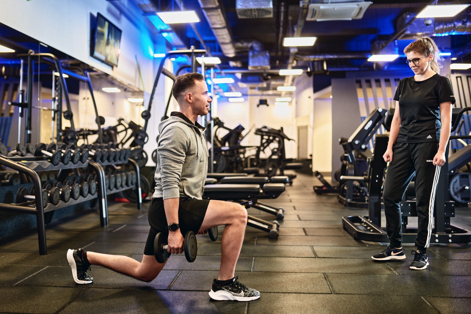 Mężczyzna w szarej bluzie wykonuje wykrok z hantlami w nowoczesnej siłowni, obserwowany przez kobietę w czarnym dresie.