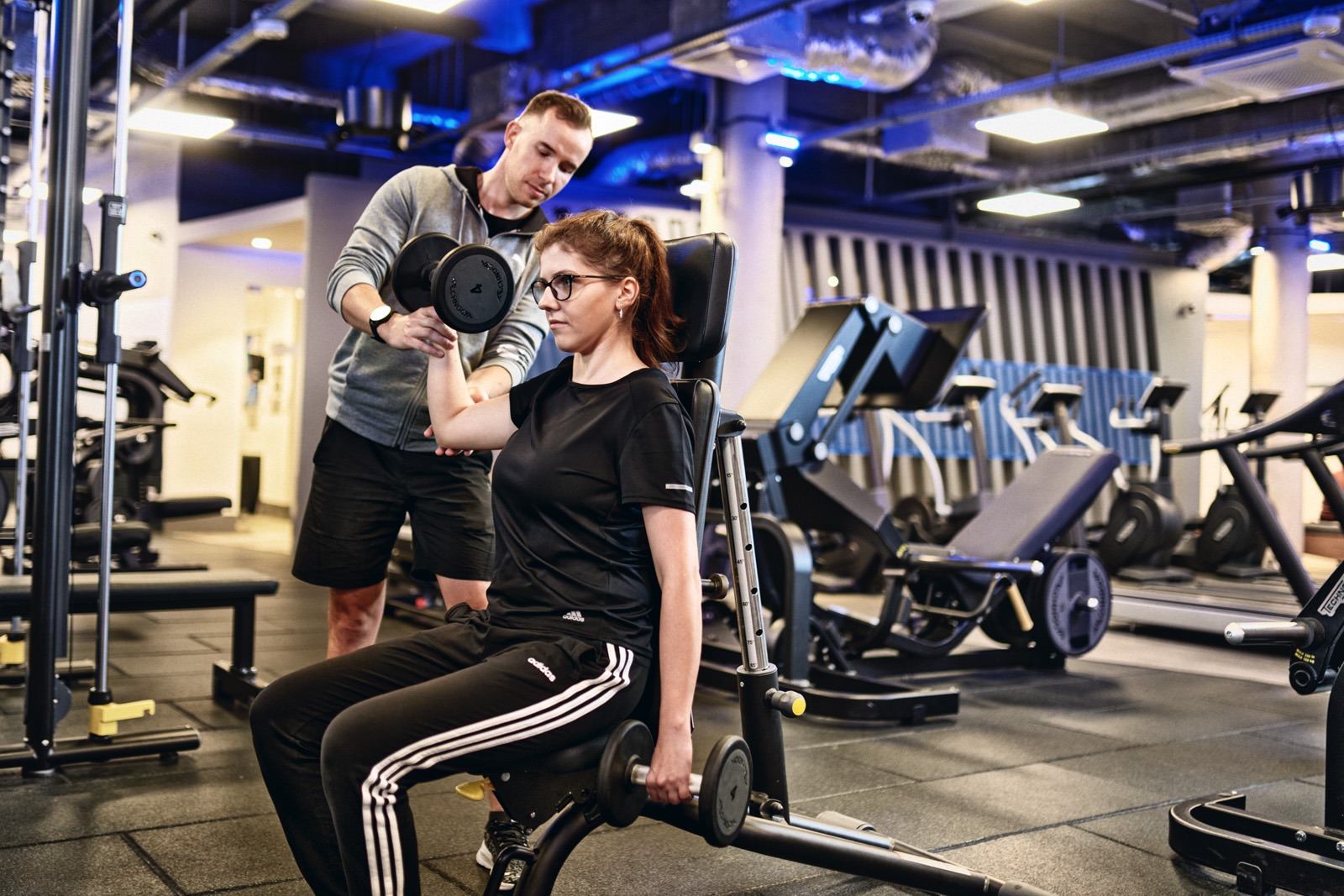 Kobieta w okularach ćwiczy biceps na maszynie w siłowni pod okiem mężczyzny z zegarkiem, który trzyma jej ramię.